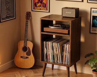 Puede incluir: Un soporte para tocadiscos de madera oscura con patas cónicas, que cuenta con un estante superior abierto con un tocadiscos y un estante inferior lleno de discos de vinilo. Un altavoz Marshall se sitúa sobre la superficie superior.