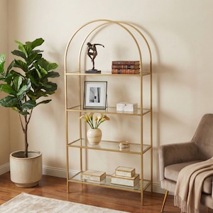 May include: A gold metal etagere with glass shelves. The arched top design adds a decorative element. The shelves display books, a framed picture, decorative boxes, and a vase of flowers. A potted plant and a chair are nearby.