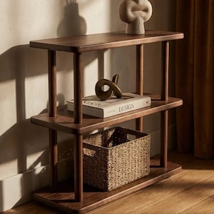 Pode incluir: Uma estante de madeira castanha escura com tr&ecirc;s n&iacute;veis. A prateleira superior tem uma escultura creme. A prateleira do meio tem um livro com a palavra "DESIGN" e uma escultura de bronze. Uma cesta de vime est&aacute; na prateleira inferior.