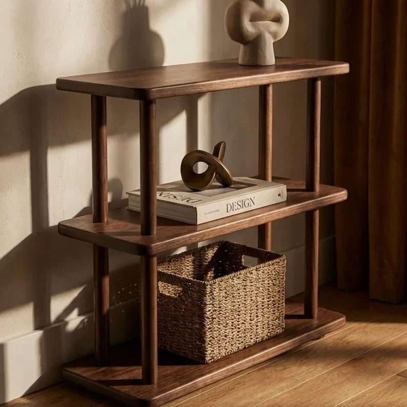 Pode incluir: Uma estante de madeira castanha escura com tr&ecirc;s n&iacute;veis. A prateleira superior tem uma escultura creme. A prateleira do meio tem um livro com a palavra "DESIGN" e uma escultura de bronze. Uma cesta de vime est&aacute; na prateleira inferior.