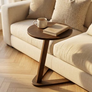 May include: A dark brown wooden side table with a circular top and a single angled support leg connected to a triangular base. A white ceramic mug and a closed beige book with visible text on the cover are placed on the table surface.