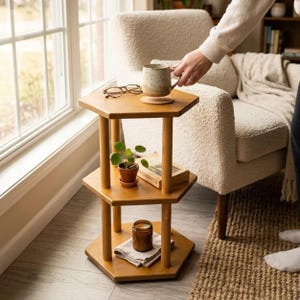 May include: A three-tiered wooden side table with a hexagonal shape. The top tier holds a speckled ceramic mug and tortoiseshell glasses. The middle tier features a small potted plant and a book. The bottom tier displays a candle and a folded cloth.