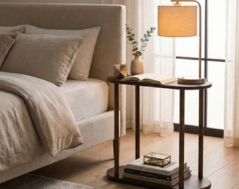 May include: A dark wood oval side table with two tiers, featuring a lit lamp with a beige drum shade, an open book, a small decorative object, and a stack of books on the lower shelf, positioned next to a beige upholstered bed.