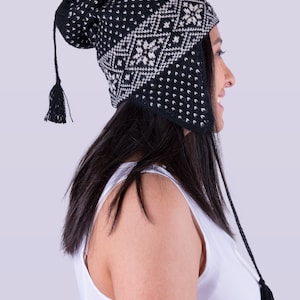 Puede incluir: Gorro de punto negro y blanco con un patrón geométrico y orejeras. El gorro tiene un largo borlón en la parte superior y un borlón más corto en cada orejera.