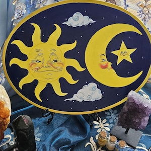 May include: Oval artwork depicting a smiling sun and moon with a star against a dark blue backdrop. The sun and moon are yellow with facial features. White clouds and gold trim complete the design.