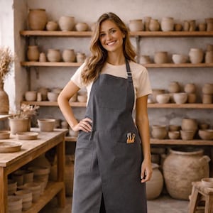Può includere: Grembiule grigio scuro con due tasche anteriori e un cinturino al collo. Il grembiule è indossato in un laboratorio di ceramica, con scaffali di ceramiche sullo sfondo. Il grembiule ha un design frontale diviso.