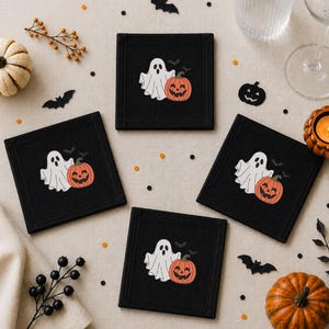 Puede incluir: Cuatro posavasos cuadrados de tela negra con un diseño bordado de un fantasma blanco que sostiene una calabaza naranja tipo linterna. Cada posavasos muestra el mismo motivo de fantasma y calabaza, acentuado por pequeños murciélagos negros.