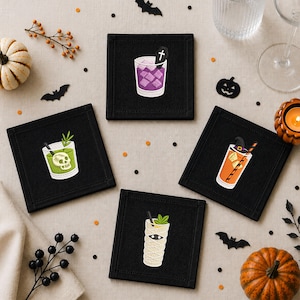 Könnte beinhalten: Ein Set aus vier quadratischen schwarzen Stoffuntersetzern, die jeweils mit einem gestickten Halloween-Cocktail-Motiv versehen sind: ein lila Getränk mit Sarg, ein grünes mit Totenkopf, ein orangefarbenes mit Hexenhut und ein weißes, mumifiziertes Getränk.