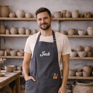 Könnte beinhalten: Eine anthrazitfarbene Schürze mit dem Namen Jack in weißer Schreibschrift auf der Brust. Die Schürze hat ein Nackenband, zwei Vordertaschen und eine kleine Seitentasche mit drei gelben Werkzeugen, getragen über einem beigen T-Shirt.