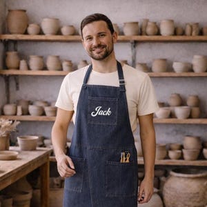 Könnte beinhalten: Eine dunkelblaue Jeansschürze mit einer Brusttasche, auf der der Name Jack in weißer Schreibschrift gestickt ist, zwei großen unteren Taschen und einem verstellbaren Nackenband. Die Schürze wird über einem cremefarbenen T-Shirt getragen.