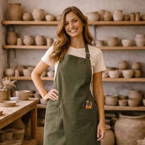 Puede incluir: Un delantal verde oliva con dos bolsillos delanteros y un diseño largo. El delantal tiene una correa que va alrededor del cuello y una hebilla. El delantal se usa sobre una camiseta blanca y unos vaqueros azules. El fondo muestra cerámica en estantes.