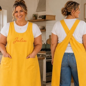 Peut inclure: Un tablier jaune moutarde avec une encolure dégagée, deux poches plaquées à l'avant et un design à dos croisé. Le devant du tablier est brodé du prénom Julia en écriture cursive. Le tablier est porté sur un t-shirt blanc et un jean bleu.