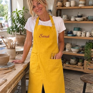 May include: A mustard yellow apron featuring a white t-shirt underneath. The apron has a square chest pocket embroidered with the name Sarah in a dark red script font, two large lower pockets, and adjustable straps with silver-toned metal hardware.