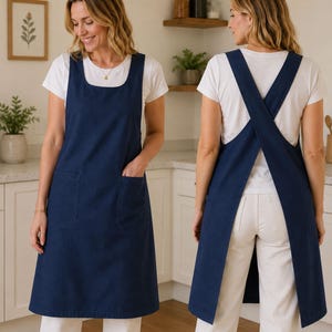 May include: A navy blue apron featuring a square neckline, two front patch pockets, and a cross-back design with wide shoulder straps. The apron is worn over a white t-shirt and white pants, showing both the front view and the back view.