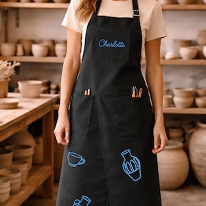 Può includere: Un grembiule nero con una tasca sul petto ricamata con il nome Charlotte in filo blu. Il grembiule include due ampie tasche in vita contenenti strumenti per ceramica e grafiche ricamate in blu raffiguranti una tazza e due vasi.