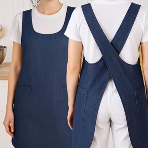May include: A dark blue denim apron with a square neckline and two front pockets. The apron has a cross-back design. The person is wearing a white t-shirt underneath.