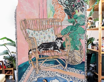 May include: Woven throw blanket featuring a whimsical illustration of two cats, one orange and one black and white, in a room with a wicker chair and potted plants. The color palette includes pink, green, and blue tones, with a fringed edge.