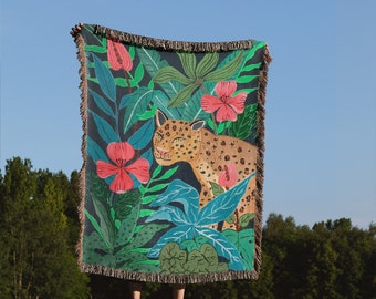Op de afbeelding: Een geweven tapijtdeken met een jungle scène met een luipaard, weelderig groen gebladerte en roze bloemen.