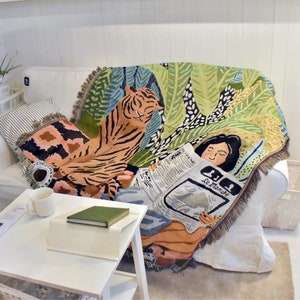 May include: A woven throw blanket with a tiger and jungle leaf design. The blanket is draped over a white sofa. A woman is depicted reading a newspaper with the text "Le Monde". The blanket has a fringed edge.
