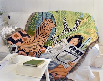 Puede incluir: Una manta tejida con un diseño de tigre y hojas de la jungla. La manta está sobre un sofá blanco. Se representa a una mujer leyendo un periódico con el texto "Le Monde". La manta tiene un borde con flecos.