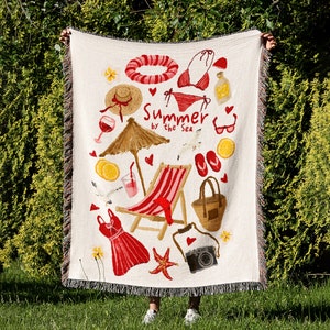 Puede incluir: Tapiz tejido blanco con temática de verano. Presenta una silla de playa a rayas rojas y blancas, un traje de baño a rayas rojas y blancas, una sombrilla de playa a rayas rojas y blancas, una bolsa de playa a rayas rojas y blancas y el texto "Summer by the Sea".