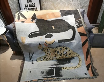 Puede incluir: Una manta tejida con ilustraciones de gatos. Uno es negro con collar, el otro es leopardo. La manta tiene un fondo azul claro, bordes con flecos y el texto "WORK HARD AND BE NICE TO PEOPLE" en el fondo.