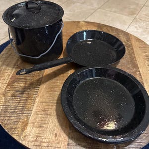 May include: A black speckled enamelware camping cookware set. The set includes a pot with a lid and handle, a frying pan with a long handle, and a plate. The cookware is arranged on a wooden surface.