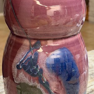 Może przedstawiać: Ręcznie wykonany ceramiczny wazon o pękatym korpusie i węższej szyi. Wazon ma bordową glazurę z abstrakcyjnymi niebieskimi, zielonymi i białymi wzorami. Podstawa jest w jasnobrązowym kolorze.