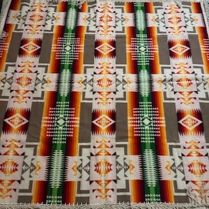 May include: A rectangular blanket with a Native American-inspired design. The blanket features a repeating pattern of geometric shapes in white, gray, orange, red, and green. The edges are finished with a fringe.