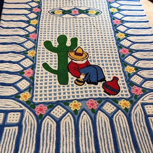 May include: A colourful, handmade rug with a Mexican theme. The rug features a central design of a green cactus and a person wearing a sombrero, with a red vase. The border is blue and white, with floral accents. The rug is approximately 152 cm long.
