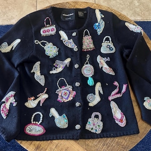May include: A black long-sleeve cardigan featuring a button-front closure and intricate beadwork designs. The embellishments include various styles of high-heeled shoes and handbags in multiple colors, including pink, blue, silver, and gold.