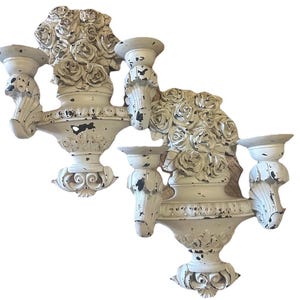 May include: Two off-white wall sconces with a distressed finish. Each sconce features a central vase overflowing with rose sculptures and three candle holders. The sconces have ornate detailing and a vintage aesthetic.