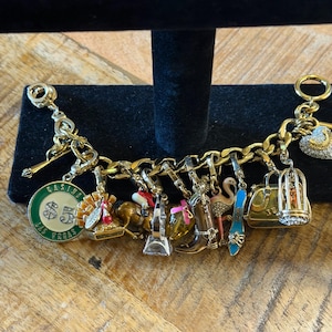 May include: Gold-toned charm bracelet featuring various charms, including a casino chip, a horse, a high heel, a flamingo, and a handbag. The charms are detailed with enamel and rhinestones. The bracelet is displayed on a black stand against a wooden background.
