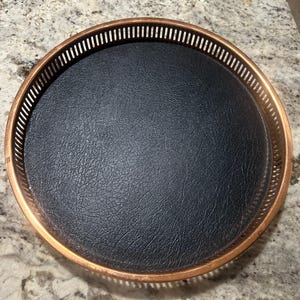 May include: A round decorative tray with a copper-colored metal rim and a black leather-like interior. The rim features a series of vertical slots. The tray is designed for serving or display.