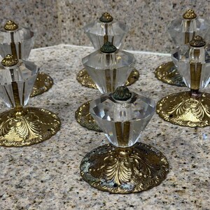 Puede incluir: Un juego de siete pomos para armarios con cabezas de cristal transparente tallado en forma de diamante, montados sobre bases metálicas circulares y ornamentadas en tono latón antiguo con grabados de hojas y un pequeño orificio.