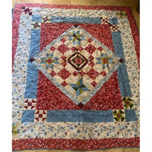 Puede incluir: Un edredón grande, hecho a mano, con un diseño de patchwork. El edredón presenta un patrón de diamante central en rojo, blanco y azul, rodeado de bordes florales y geométricos. Sus dimensiones son de aproximadamente 203 cm por 229 cm.