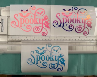 May include: Three "Spooky" vinyl decals in pink, purple, and blue gradients, featuring bats, ghosts, and skulls. The decals are on a white background, with a ruler and paper cutter visible.