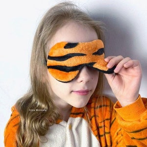 Puede incluir: Un niño que lleva un mono de estampado de tigre sostiene una máscara para dormir de estampado de tigre sobre sus ojos. La máscara está hecha de tela suave y afelpada y tiene rayas negras sobre un fondo naranja.