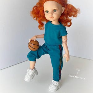 May include: A doll with red curly hair wearing a teal blue tracksuit with white trainers and holding an orange basketball. The tracksuit has the words "GENIUS NICE NICE NICE" printed on the side.