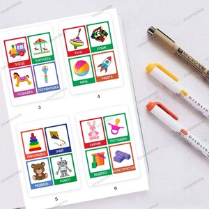 May include: A set of colorful flashcards with pictures of toys and their names in Russian. The cards are arranged in rows of four, with each card featuring a different toy. The toys include a train, a carousel, a spinning top, a building block, a ball, a rocket, a rocking horse, a rattle, a pyramid, a kite, a bunny, a pacifier, a bucket, a toy robot, a teddy bear, and a set of building blocks.