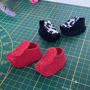 May include: Four felt doll shoes. Two are red and two are black with a white and black animal print. The shoes are on a green cutting mat with white markings.