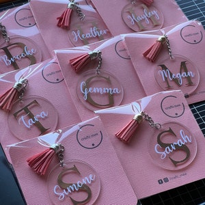 May include: A set of ten pink and gold personalised keychains with tassels. Each keychain features a clear acrylic circle with a gold letter and a name written in white. The names are Brooke, Tara, Gemma, Heather, Hayley, Megan, Simone, and Sarah.