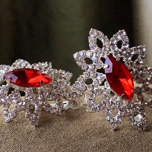 May include: Two silver napkin rings with a red rhinestone in the center of each. The rings are decorated with clear rhinestones in a floral design.