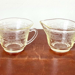 May include: A set of two vintage glass sugar and creamer bowls with a delicate floral pattern. The bowls are clear with a pale yellow tint and have two handles each.
