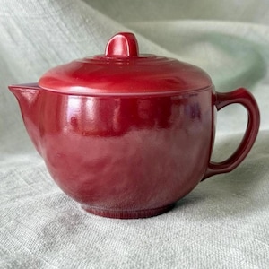 May include: A red ceramic teapot with a lid and handle. The teapot has a rounded body, a spout, and a curved handle. The lid has a small knob on top. The teapot is sitting on a light-colored surface.
