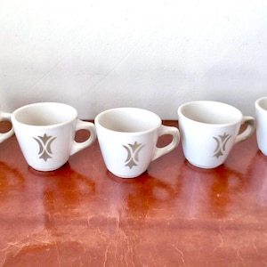 May include: Set of five white ceramic coffee cups with a gray stylized design. The cups have a rounded shape and a handle on the right side.
