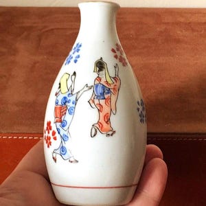 May include: A white ceramic sake bottle with a floral design and two women in traditional Japanese clothing painted on the side.