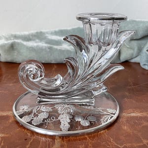 May include: A clear glass candlestick with a grapevine design. The candlestick has a single candle holder and a base with a silver-toned grapevine design.