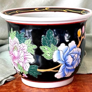 May include: Black ceramic planter with a white rim and floral design. The planter features pink and blue flowers with green leaves. The rim has a red and gold accent. A decorative piece for indoor or outdoor use.