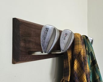 May include: A wooden wall-mounted rack with two silver golf wedges hanging from it. The wedges are labeled "Warrior" and "Staff". A plaid shirt is hanging on the rack.